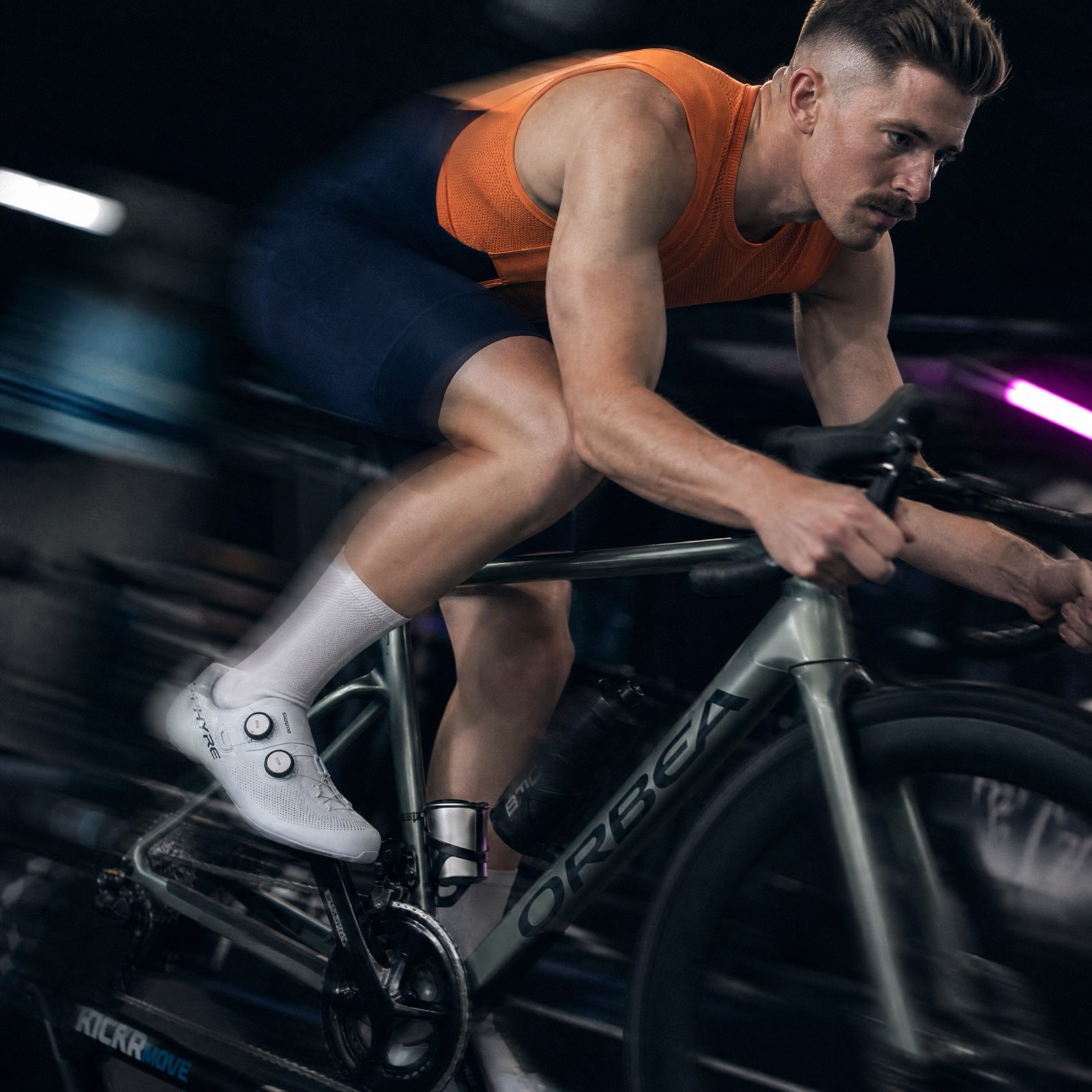 A rider in an orange top on an indoor trainer, the room dark behind him.
